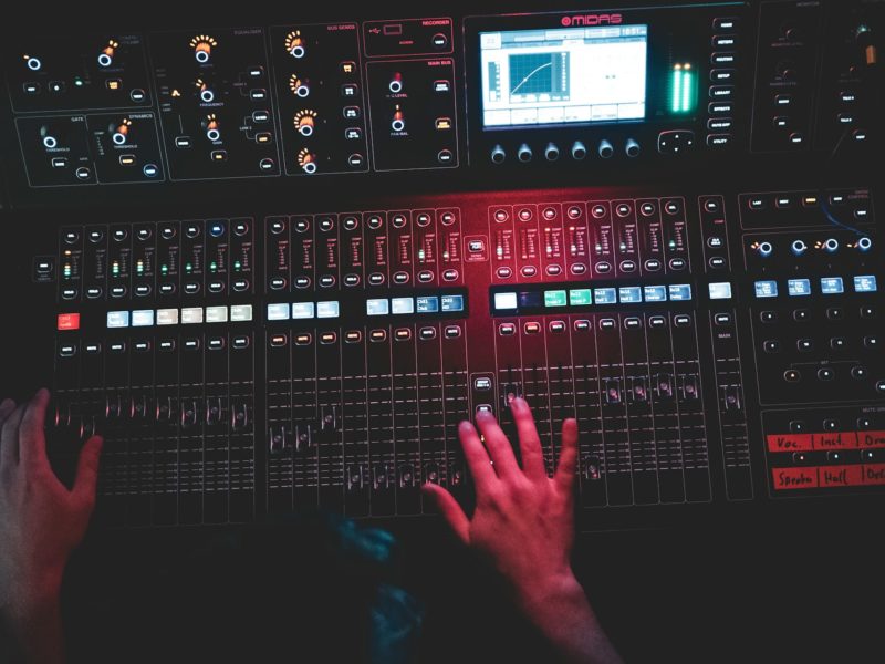 Ein Mensch bedient ein professionelles Audiomischpult mit zahlreichen Schiebereglern, Tasten, Drehknöpfen und einem digitalen Display. Die Szene ist dunkel und in rotes Licht getaucht, wodurch eine atmosphärische Studio- oder Live-Umgebung entsteht.