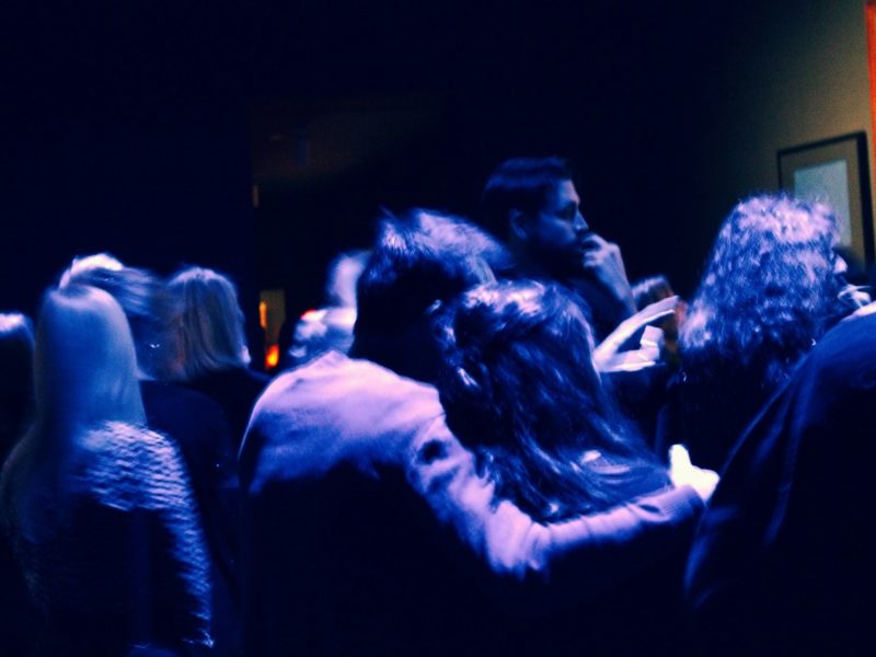 Eine Gruppe von Menschen steht dicht gedrängt in einem dunklen Raum mit schwacher, bläulicher Beleuchtung. Einige Personen scheinen sich zu unterhalten, während ein Paar sich innig umarmt. Im Hintergrund sind verschwommene Umrisse von Bildern an der Wand zu erkennen, eines davon zeigt eine Figur im Sonnenlicht. Die Szene wirkt wie auf einer Party oder bei einer Kunstausstellung, leicht verschwommen durch Bewegung.