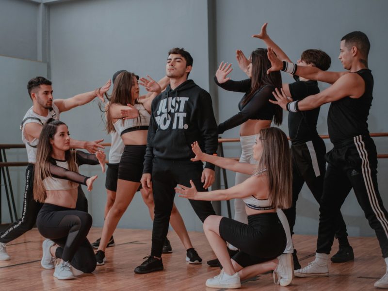 Eine Tanzgruppe aus acht jungen Erwachsenen führt eine choreografierte Pose in einem Tanzstudio auf. In der Mitte steht ein Mann mit schwarzem Hoodie, auf dem „JUST DO IT.“ steht, während die anderen ihn mit ausgestreckten Armen und dramatischen Gesten umkreisen. Die Szene wirkt dynamisch und ausdrucksstark, typisch für eine moderne Tanzaufführung oder Probe.