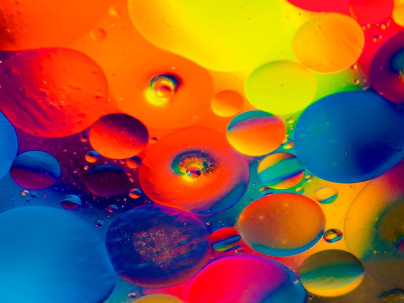 Bunte Flüssigkeitstropfen in leuchtenden Farben wie Blau, Rot, Gelb, Orange und Pink schweben nebeneinander und übereinander in einer abstrakten Komposition. Die Formen erinnern an Öl- oder Wassertropfen unter einem Mikroskop, die Licht in regenbogenartigen Farben reflektieren. Das Bild wirkt lebendig und künstlerisch.