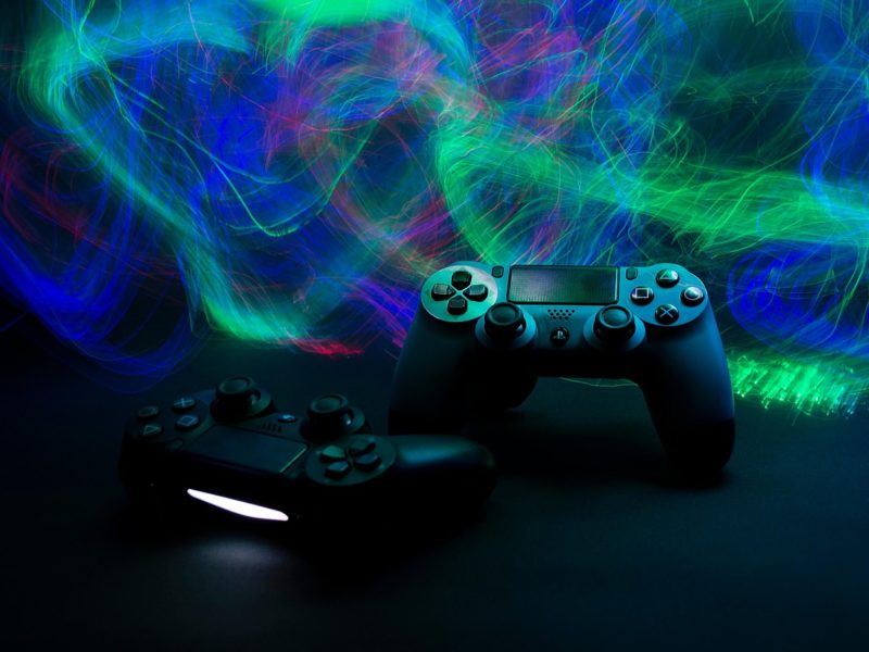 Zwei PlayStation-Controller liegen auf einer dunklen Oberfläche, umgeben von leuchtenden, abstrakten Lichtspuren in Blau, Grün und Rot, die eine dynamische und futuristische Atmosphäre erzeugen.