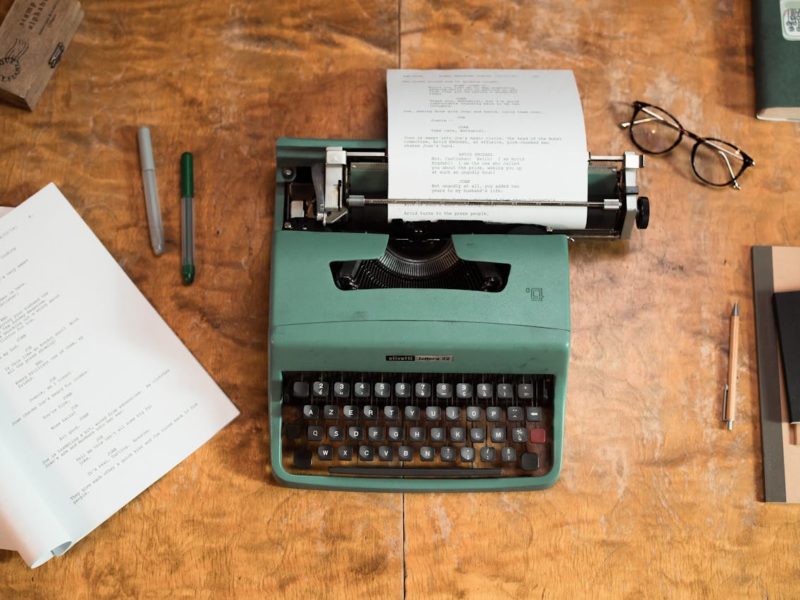 Eine grüne Schreibmaschine (Olivetti Lettera 32) steht auf einem hölzernen Schreibtisch. In der Maschine steckt ein getipptes Drehbuch oder Manuskript. Um die Schreibmaschine liegen zwei Stifte, ein aufgeklapptes Skript, ein Notizblock mit Stift, eine Brille und ein Buch. Die Szene wirkt nostalgisch und kreativ, als wäre jemand gerade beim Schreiben.