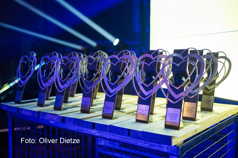 Mehrere Trophäen mit herzförmigem Design stehen in Reihen auf einem Tischgitter. Jede Auszeichnung hat einen rechteckigen Sockel mit einer kleinen Plakette und enthält eine Kugel im Zentrum des Herzens. Die Szene ist in blaues und gelbes Bühnenlicht getaucht.