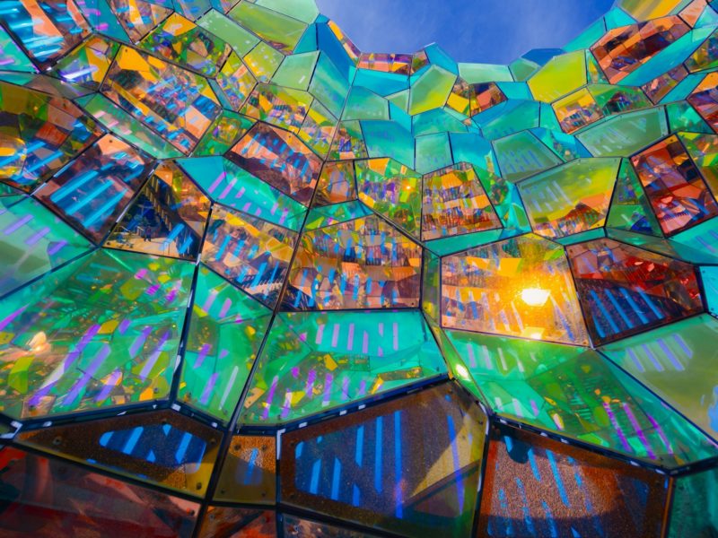 Bunte, facettenreiche Struktur aus transparenten, geometrischen Glaspaneelen in leuchtenden Farben wie Türkis, Gelb, Violett und Orange; das Licht bricht sich darin spektakulär und erzeugt lebendige Reflexionen und Muster vor einem blauen Himmel.