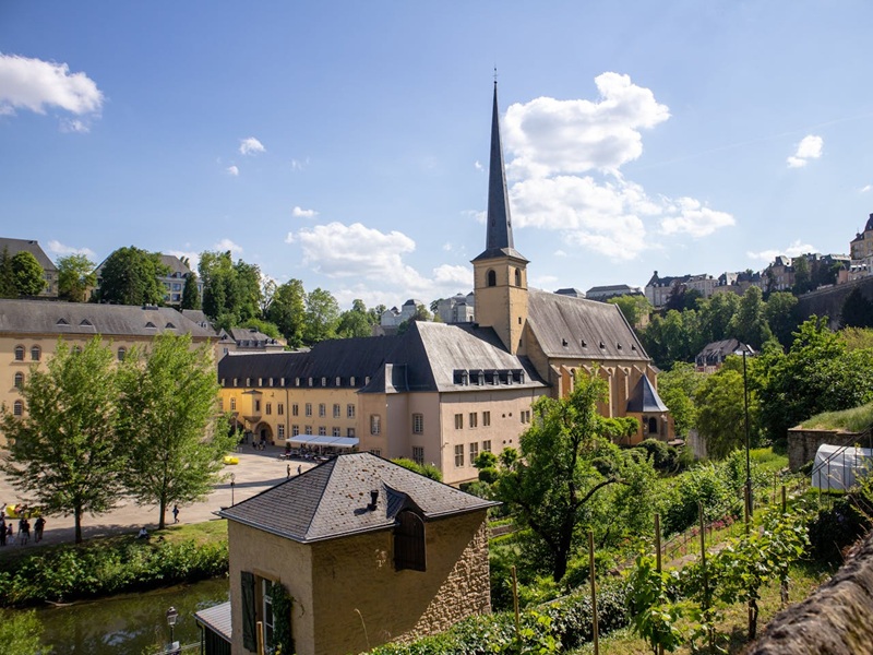 Historisches Gebäudeensemble mit einer großen Kirche und spitzem Turm in Luxemburg-Stadt, umgeben von Bäumen und alten Gemäuern an einem sonnigen Tag mit blauem Himmel und vereinzelten Wolken. Im Vordergrund ein kleiner Fluss und Gärten mit Weinreben.