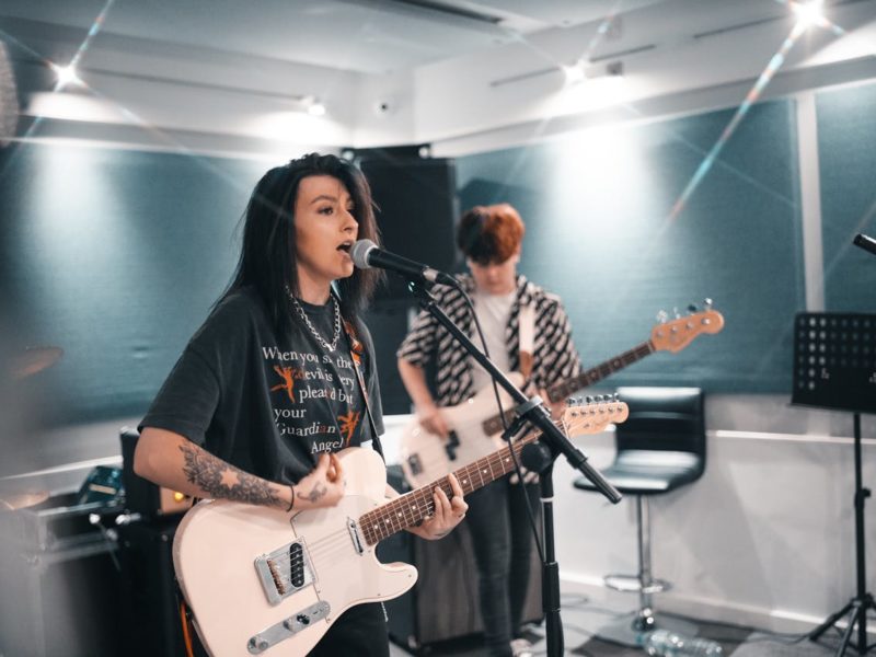 Zwei junge Musiker:innen in einem Proberaum. Im Vordergrund steht eine Person mit dunklen Haaren und Tätowierungen, die in ein Mikrofon singt und eine E-Gitarre spielt. Im Hintergrund spielt eine weitere Person Bassgitarre. Der Raum ist modern mit schallgedämpften Wänden ausgestattet, mehrere Mikrofone und Verstärker sind sichtbar.