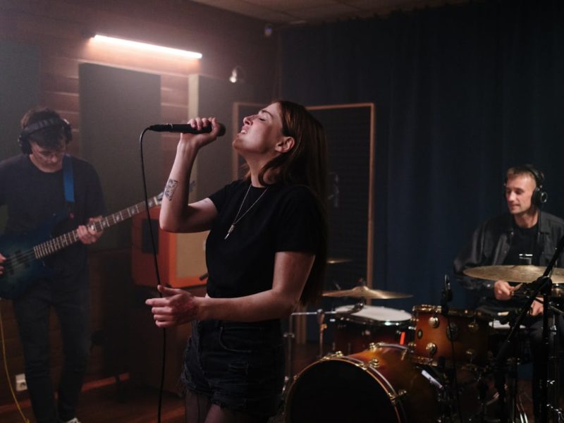 Drei Musiker proben in einem Tonstudio – eine Sängerin im Vordergrund singt leidenschaftlich ins Mikrofon, während links ein Bassist und rechts ein Schlagzeuger spielen.