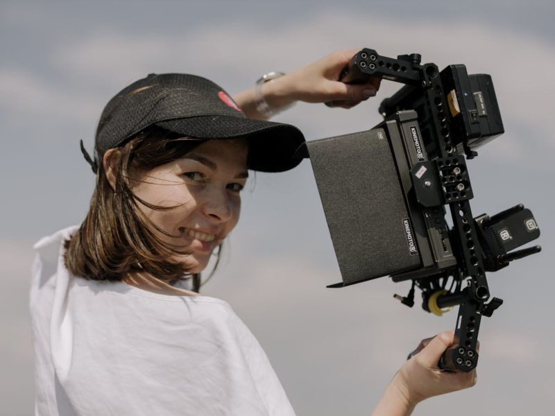 Eine junge Frau trägt ein schwarzes Basecap und hält eine große, professionelle Filmkamera über ihrer Schulter. Sie lächelt in die Kamera, während der Himmel im Hintergrund leicht bewölkt ist.