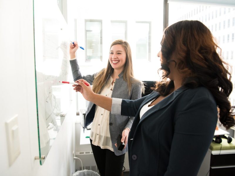 Zwei Frauen in Business-Kleidung stehen vor einem Whiteboard in einem hellen Büro. Eine Frau hält einen blauen Marker und lächelt, während die andere mit einem roten Marker auf das Board zeigt. Beide wirken konzentriert und engagiert im Gespräch.