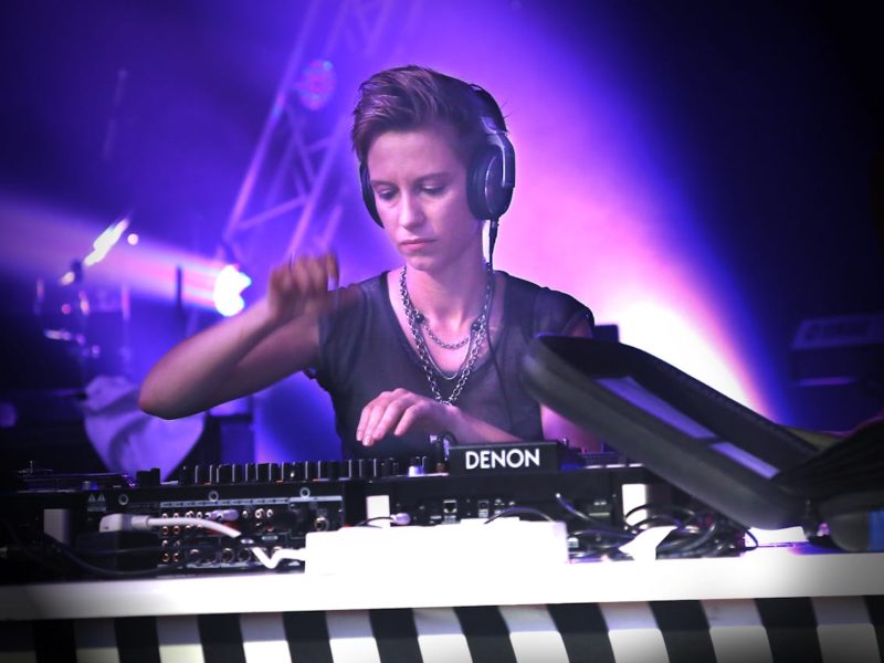 DJ mit kurzen Haaren und Kopfhörern steht an einem Mischpult der Marke Denon. Die Person trägt eine schwarze, durchsichtige Oberbekleidung und Ketten, konzentriert sich auf das Auflegen und ist von violetten Bühnenlichtern umgeben. Im Vordergrund ist ein gestreifter Tischrand sichtbar.
