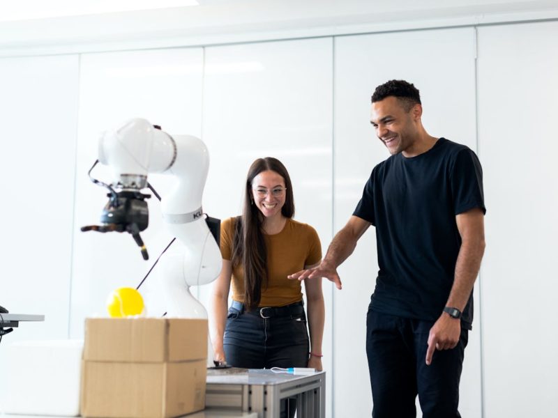 Zwei junge Erwachsene stehen lachend vor einem weißen Roboterarm in einem hellen Raum. Der Roboter bewegt einen gelben Gegenstand über einen Tisch mit einem Karton, während die beiden aufmerksam zusehen und begeistert wirken.