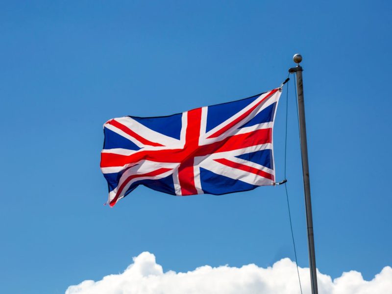 Auf dem Bild ist die Flagge des Vereinigten Königreichs (Union Jack) zu sehen, die an einem Fahnenmast im Wind weht. Der Himmel im Hintergrund ist klar blau mit einigen weißen Wolken am unteren Bildrand.