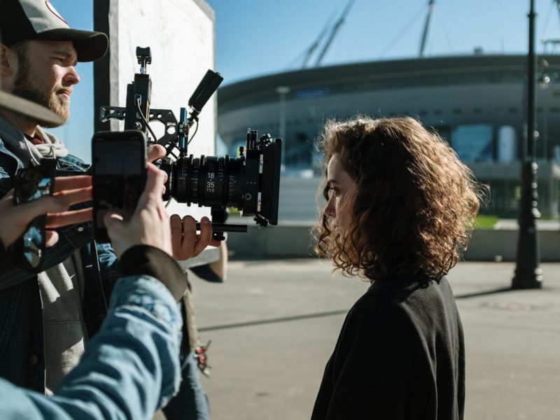 Eine Filmszene wird im Freien gedreht. Ein Kameramann mit Kappe hält eine professionelle Filmkamera auf eine Frau mit lockigen Haaren, die im Profil zu sehen ist. Neben der Kamera ist eine weitere Person mit Smartphone sichtbar. Im Hintergrund steht ein modernes Stadion.