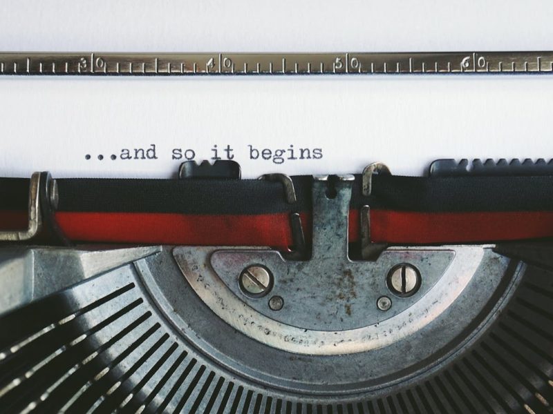 Nahaufnahme einer Schreibmaschine, die auf ein Blatt Papier den Satz „...and so it begins“ tippt.