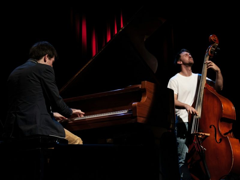 Zwei Musiker auf einer Bühne, einer spielt Klavier, der andere Kontrabass. Beide sind konzentriert und in die Musik vertieft, die Szene ist dunkel mit rotem Bühnenlicht im Hintergrund.
