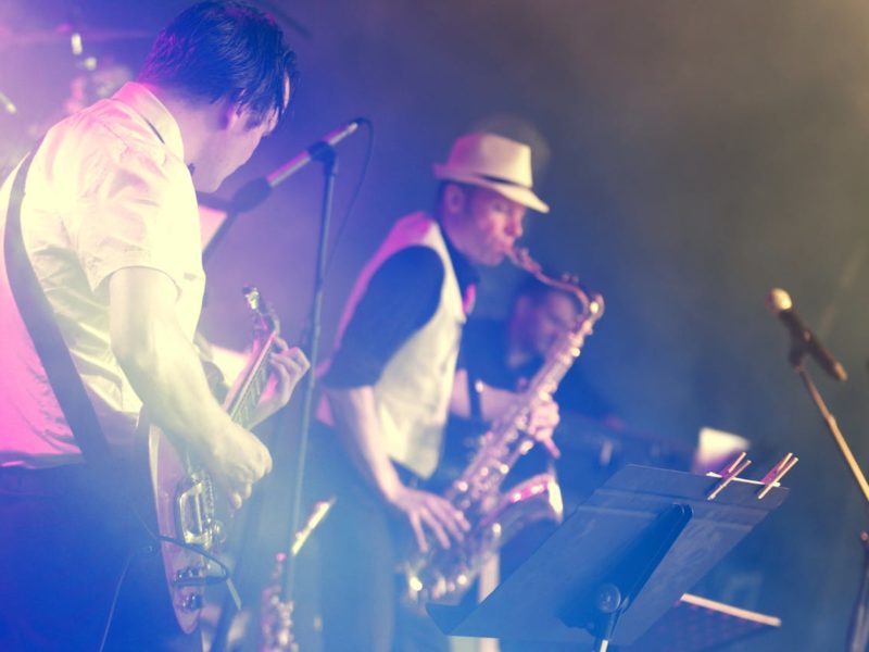 Zwei Musiker auf einer Bühne in atmosphärischer blau-violetter Beleuchtung mit Nebel-Effekten. Im Vordergrund spielt ein Gitarrist im weißen Hemd eine E-Gitarre, während im Hintergrund ein Saxophonist mit Fedora-Hut musiziert. Unten rechts ist ein Notenständer mit Notenblättern sichtbar.