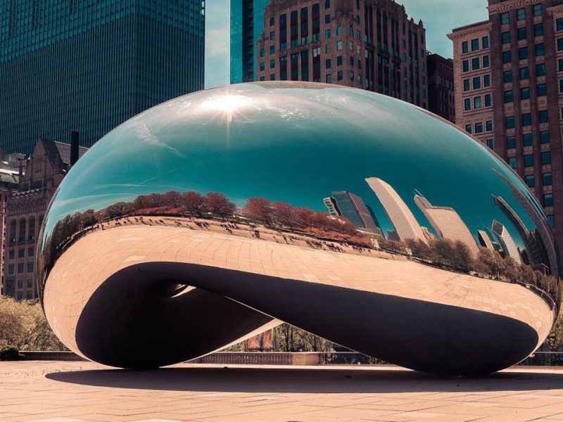 Glänzende, bohnenförmige Skulptur „Cloud Gate“ in Chicago, die die Skyline und den Himmel in ihrer spiegelnden Oberfläche reflektiert, umgeben von hohen Gebäuden und Bäumen.
