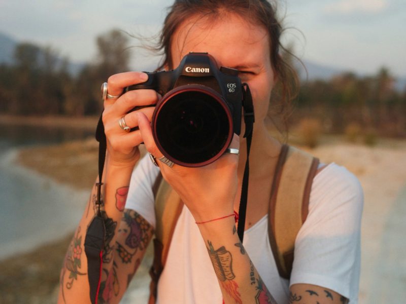 Person mit tätowierten Armen und weißem T-Shirt hält eine Canon-Kamera vor das Gesicht und macht ein Foto im Freien, mit unscharfem Hintergrund aus Wasser, Bäumen und Bergen bei warmem Licht.