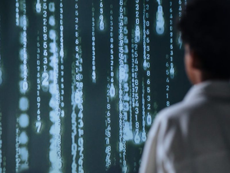 Eine Person steht vor einem großen Bildschirm oder einer Projektion, auf der in grünlich-weißer Farbe Zahlen vertikal von oben nach unten laufen. Die Szene erinnert an digitale Codes oder Datenströme und vermittelt eine technologische, futuristische Atmosphäre. Die Person ist von hinten zu sehen und trägt ein helles Oberteil.