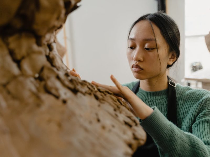 In einem Atelierraum formt eine junge Person mit langen, dunklen Haaren eine große Skulptur aus Ton oder Lehm. Sie steht sehr nah am Werkstück und berührt die Oberfläche vorsichtig mit beiden Händen, sichtbar vertieft in die Arbeit. Das Licht fällt weich durch ein Fenster im Hintergrund, und die Umgebung wirkt wie ein Künstler- oder Werkstudio.