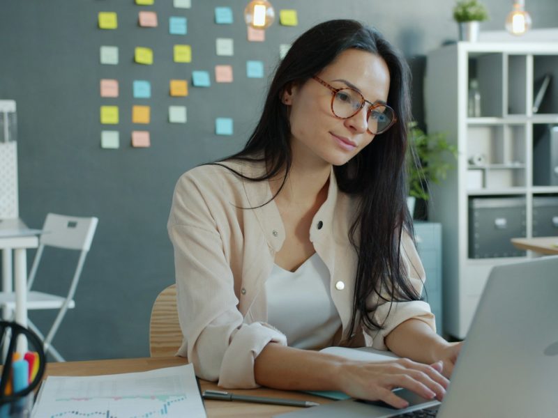 Die Szene zeigt eine Person, die konzentriert an einem Laptop arbeitet. Sie sitzt an einem ordentlich eingerichteten Schreibtisch, auf dem einige Unterlagen mit Diagrammen sowie Büro-Utensilien liegen. Im Hintergrund hängen bunte Haftnotizen an einer dunklen Wand, was nach einem kreativen oder projektorientierten Arbeitsumfeld aussieht. Regale mit Ordnern, Pflanzen und Schreibtischlampen ergänzen den Raum und verleihen ihm eine moderne, strukturierte Atmosphäre. Insgesamt wirkt das Bild wie eine ruhige, produktive Bürosituation – jemand, der fokussiert an einer Aufgabe oder einem Projekt sitzt.