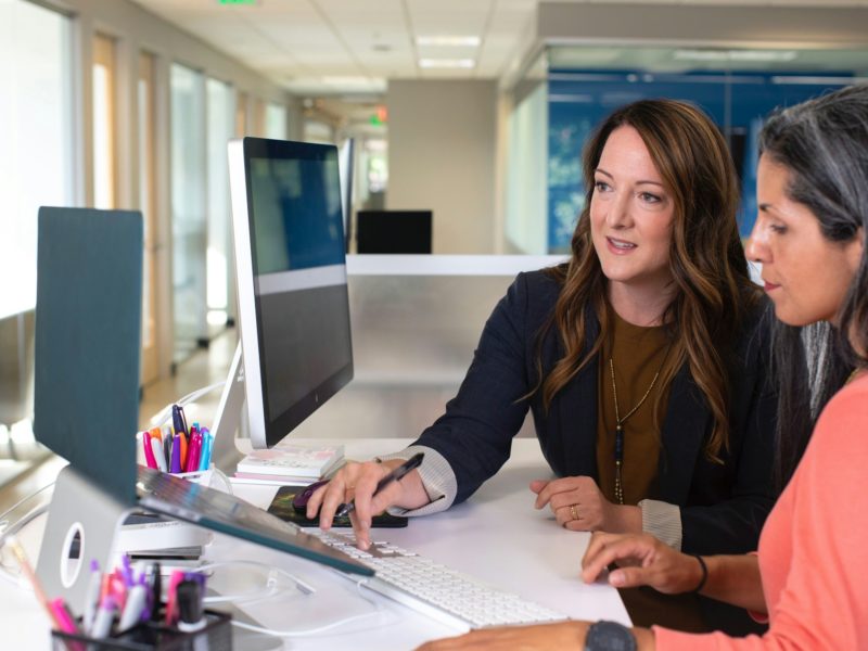 Auf dem Bild sind zwei Personen in einem modernen Büro zu sehen. Sie sitzen gemeinsam an einem großen Computerbildschirm und arbeiten konzentriert an etwas. Die Person links zeigt mit einem Stift auf den Monitor, offenbar um etwas zu erklären oder gemeinsam zu besprechen. Beide wirken engagiert und fokussiert. Auf dem Schreibtisch stehen bunte Stifte, Arbeitsmaterialien und eine Tastatur. Der Hintergrund zeigt einen hellen Büroflur mit Glastüren und viel Tageslicht – insgesamt eine professionelle, kollaborative Arbeitssituation.