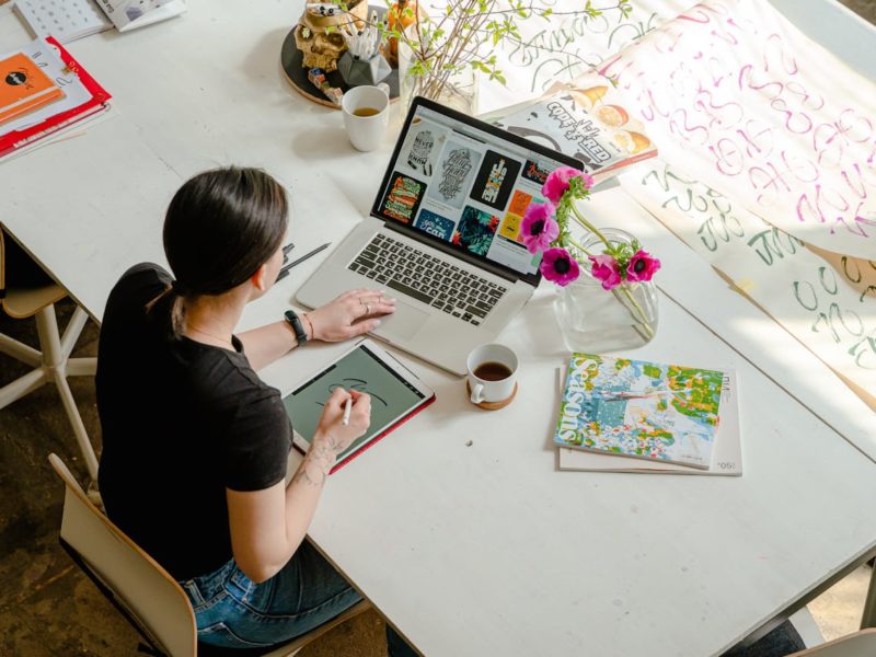 Hier ist ein Alt-Text für das Bild: "Vogelperspektive auf eine kreative Designerin an einem großen weißen Arbeitstisch. Sie arbeitet mit einem Stylus auf einem Grafiktablet, während vor ihr ein Laptop mit Designentwürfen geöffnet ist. Auf dem Tisch befinden sich eine Tasse Tee, pinke Mohnblumen in einer Vase, Magazine, Notizbücher und große Papierbögen mit handgeschriebenen Schriftzügen in verschiedenen Farben. Die Szene zeigt einen typischen kreativen Arbeitsplatz.