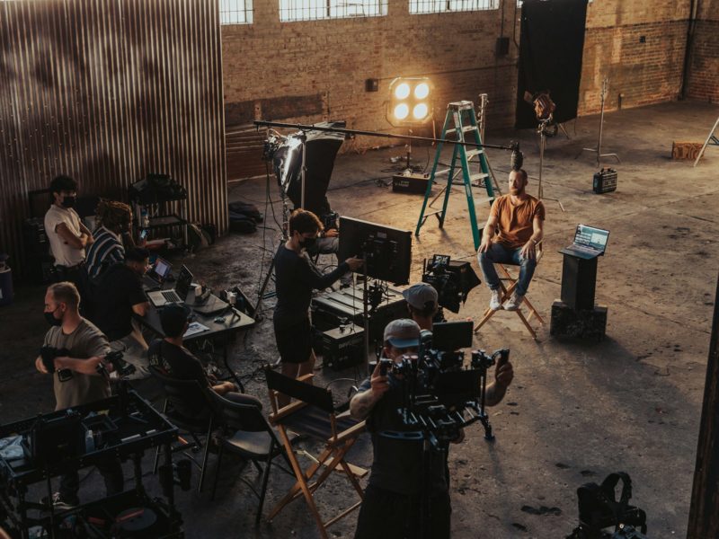 "Filmcrew bei Dreharbeiten in einer industriellen Lagerhalle mit Backsteinwänden. Ein Kameramann filmt eine Person, die auf einem Regiestuhl sitzt, während Crewmitglieder verschiedene Ausrüstung bedienen, darunter Monitore, Scheinwerfer und eine Leiter. Professionelles Videoproduktions-Setup mit mehreren koordiniert arbeitenden Teammitgliedern