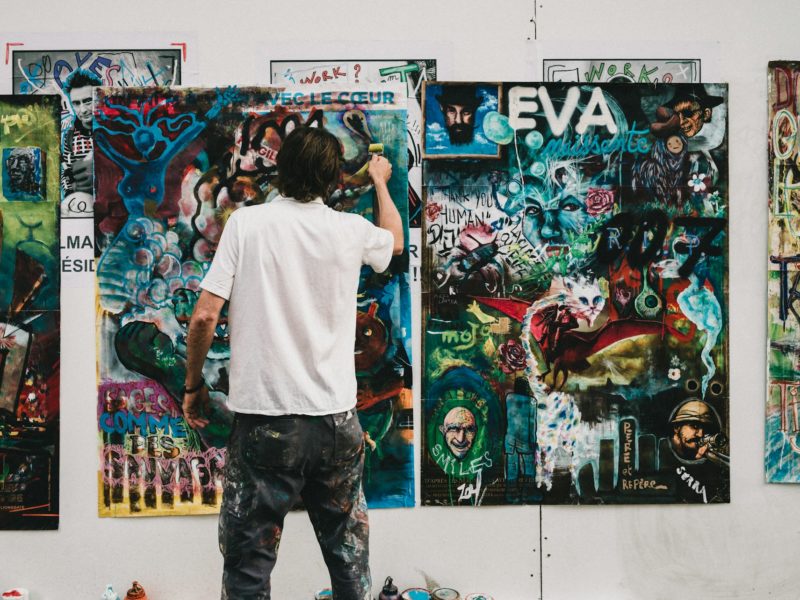 Ein Künstler in weißem T-Shirt und farbverschmierter Hose steht von hinten fotografiert vor einer weißen Wand, an der mehrere großformatige Collagen und Mixed-Media-Kunstwerke hängen. Er arbeitet gerade an einem der bunten, layerten Werke, die Textelemente wie 'EVA', 'AVEC LE CŒUR' und verschiedene Graffiti-Stile, Porträts und abstrakte Elemente in leuchtenden Farben – vor allem Blau-, Grün- und Rottönen – kombinieren. Seine Kleidung zeigt Farbspuren seiner künstlerischen Arbeit.