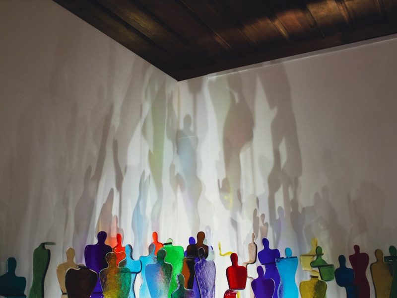 Kunstinstallation in einem Raum mit dunkler Holzdecke: Eine Reihe bunter, durchscheinender Glasfiguren in Menschenform steht vor einer weißen Wand. Die Figuren in verschiedenen Farben – Blau, Grün, Rot, Gelb, Orange und Lila – werden von hinten beleuchtet, wodurch ihre vergrößerten, verschwommenen Schatten an die Wand projiziert werden. Die Schatten überlagern sich und erzeugen ein ätherisches, traumhaftes Schattenspiel.