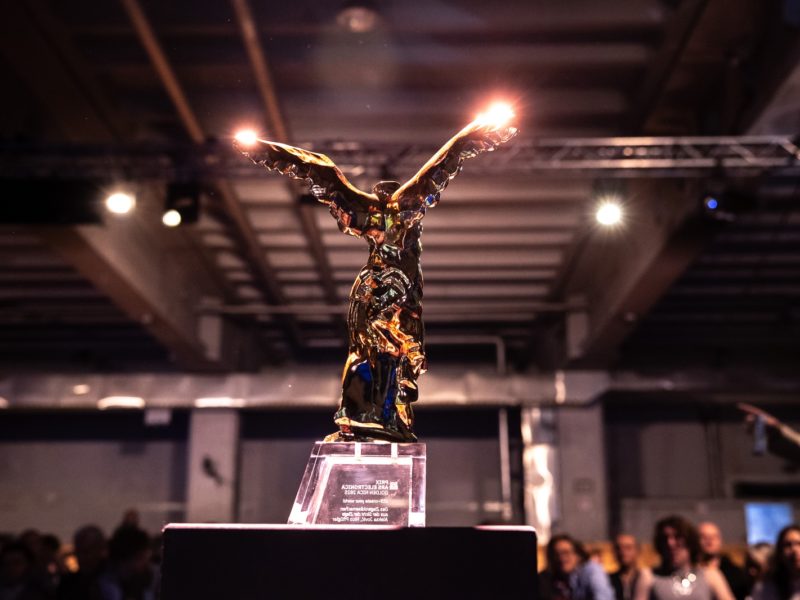Eine goldene Trophäe in Form einer Siegesgöttin mit ausgebreiteten Flügeln steht auf einem transparenten Kristallsockel. Die Statue wird dramatisch von oben beleuchtet, wodurch die Flügelspitzen hell erstrahlen. Im unscharfen Hintergrund ist eine Veranstaltungshalle mit Industriedecke und Publikum zu erkennen. Die Atmosphäre wirkt festlich und feierlich, wie bei einer Preisverleihung.