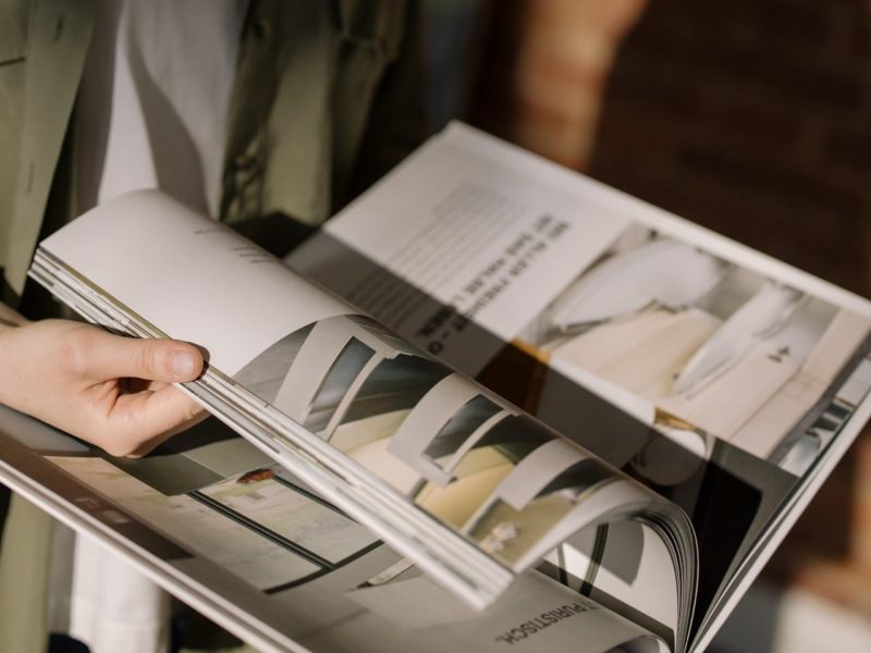 Nahaufnahme einer Person, die in einem hochwertigen Magazin oder Katalog blättert. Die Hand hält die Seiten beim Umblättern, wobei mehrere Seiten gleichzeitig in einer geschwungenen Bewegung zu sehen sind. Die Publikation zeigt Innenarchitektur- oder Designfotografien in gedeckten, neutralen Farbtönen. Im unscharfen Hintergrund trägt die Person eine olivgrüne Jacke.