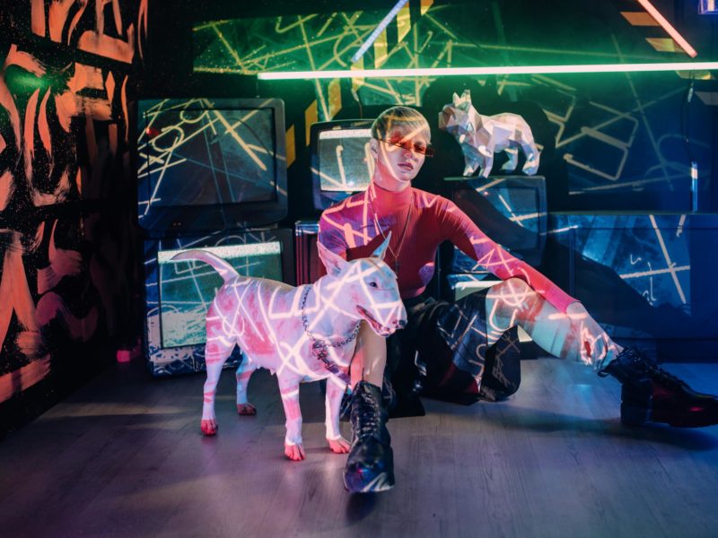 Futuristische Szene mit einer Frau in pinkem Rollkragenpullover und Sonnenbrille, die auf dem Boden neben einem weißen Bullterrier sitzt. Die beiden werden von bunten Neonlichtern in Pink, Grün und Blau beleuchtet. Im Hintergrund eine urbane Kulisse mit Graffiti-Wand in Orange und Grün, Computermonitoren und einer polygonalen Elefantenskulptur. Die Atmosphäre wirkt cyberpunk-artig und künstlerisch inszeniert.