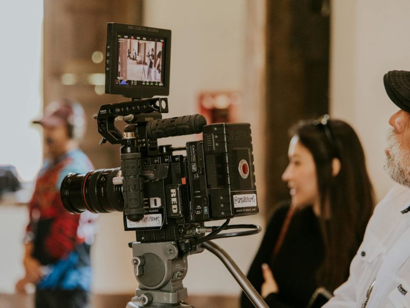 Nahaufnahme einer professionellen RED-Filmkamera auf einem Stativ am Filmset. Auf dem Monitor der Kamera ist eine Szene mit Personen zu sehen. Im unscharfen Hintergrund sind Teammitglieder erkennbar, rechts im Bild ein Mann mit grauer Baseballkappe und Bart, der die Aufnahme beobachtet.